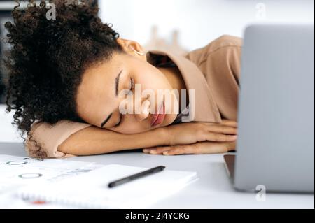 Gros plan d'une fille africaine américaine fatiguée, une femme indépendante ou étudiante, s'est endormi sur le bureau près de l'ordinateur portable, a surtravaillé de travailler ou d'étudier en ligne, a besoin de repos et de sommeil Banque D'Images