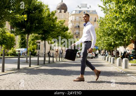 Beau jeune homme d'affaires afro-américain tenant un téléphone portable tout en traversant une rue Banque D'Images