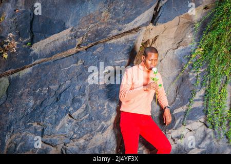 Homme vous manquant. Vêtu d'un chandail orange clair avec col haut, pantalon rouge, portant montre-bracelet, un jeune homme noir est debout contre les rochers avec lo Banque D'Images