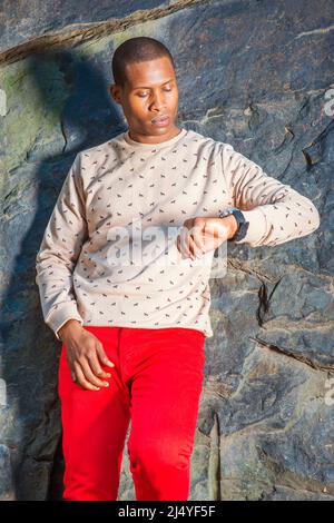 Homme qui vous attend. Robe en crème, chandail à motifs, sans collerettes, pantalon rouge, portant montre-bracelet, un jeune homme noir se penche contre les rochers, le raiis Banque D'Images