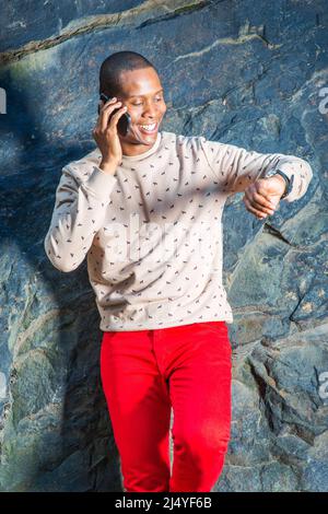 Homme qui vous attend. Robe en crème, chandail à motifs, sans collerettes, pantalon rouge, portant montre-bracelet, un jeune homme est debout près des rochers, souriant, makin Banque D'Images