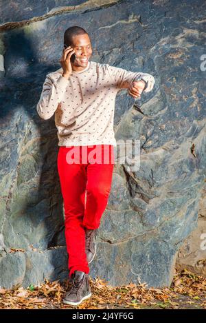 Heure. Robe de chambre crème, à motifs, chandail sans collerre, jeans rouges, chaussures de marche, portant une montre-bracelet, un jeune homme noir est debout près des rochers, souriant, ma Banque D'Images