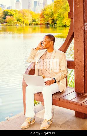 Homme d'affaires travaillant. Déguisements en blazer crème, chandail sans collerre, pantalon blanc, baskets, un jeune homme noir est assis près d'un lac dans une grande ville, smili Banque D'Images