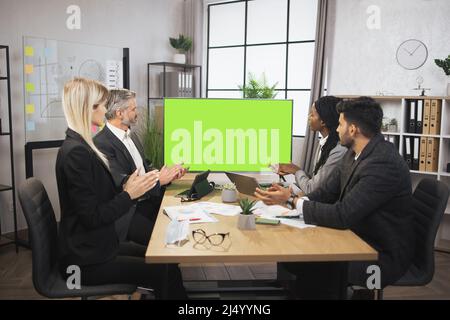 Un groupe heureux de quatre hommes d'affaires multiethniques se claquant les mains, assis à la table dans un bureau moderne, devant un immense écran de télévision plasma avec espace clé vert chromé pour le texte. Banque D'Images