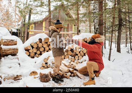 Jeune père en vêtements d'hiver assis sur des squats par pile de bois couverte de neige et passant du bois de chauffage à son fils contre leur maison de campagne Banque D'Images