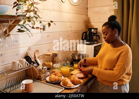 Jeune femme blaxck contemporaine en chandail tricoté jaune préparant des sandwichs avec du fromage, du jambon et des tomates fraîches hachées Banque D'Images