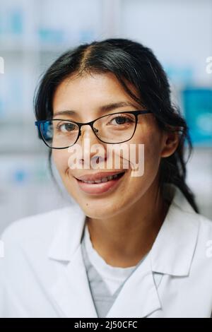 Jeune femme hispanique à succès biochimiste en lunettes et en whitecoat regardant la caméra avec le sourire tout en travaillant en laboratoire Banque D'Images