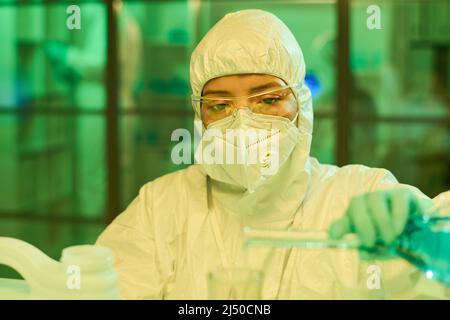 Jeune femme scientifique contemporaine dans des combinaisons de protection, des gants et un respirateur versant du liquide bleu du flacon dans d'autres verres Banque D'Images