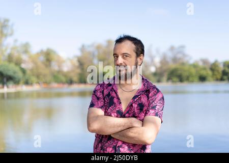 Homme mûr barbu portant une chemise fuchsia, bras croisés, regardant la caméra sur un lac Banque D'Images