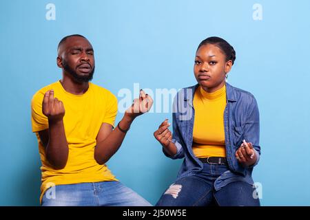 Couple faisant le geste d'argent et exigeant des récompenses d'argent pendant qu'ils sont assis dans le studio. Homme et femme se frottant les doigts pour demander le paiement, se sentant optimiste. Personnes utilisant le langage corporel pour recevoir des revenus. Banque D'Images