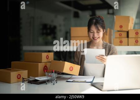 Jeune femme propriétaire de petite entreprise achats en ligne à la maison. Vérifier les commandes de clients avec la tablette numérique préparant le produit de paquet sur fond Banque D'Images