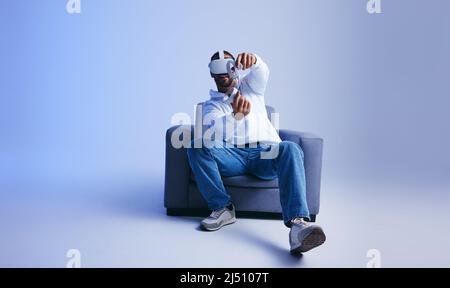 Jeune homme heureux jouant un jeu de réalité virtuelle immersif sur un canapé. Homme jouant avec des lunettes de réalité virtuelle et des contrôleurs. Joyeux jeune homme havi Banque D'Images
