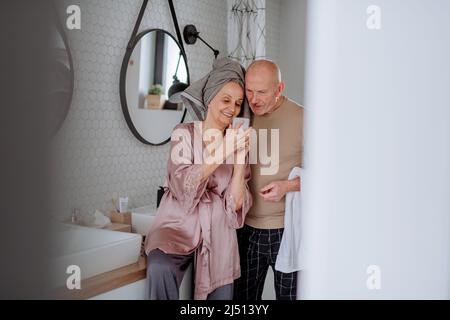 Couple senior amoureux dans la salle de bains, utilisant un smartphone, concept de routine du matin. Banque D'Images