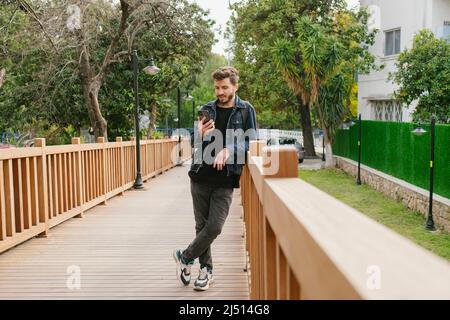 Prise de vue en longueur d'un jeune homme portant des vêtements en denim et regardant au téléphone avec des sourires. Penchée à une clôture en bois. Il attend sa petite amie. Seul homme, TH Banque D'Images