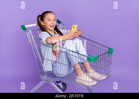 Photo de la petite écolière très impressionnée habillé jeans chemise conduite plateau shopping dactylographie gadget isolé violet couleur fond Banque D'Images