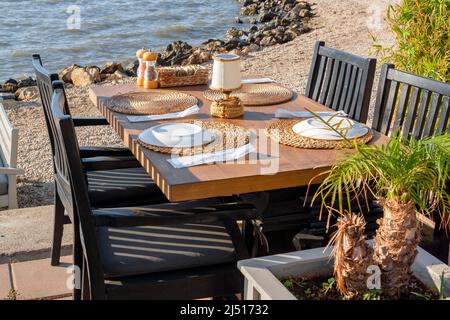 Une table et des chaises en bois avec des plats au café sur la mer attendent les clients. Petit palmier en pot. Photo horizontale. Banque D'Images