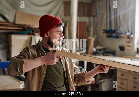 Jeune homme de charpentier caucasien avec barbe sur le visage portant une casquette tricotée rouge regardant la surface de la planche en bois pour vérifier sa qualité Banque D'Images