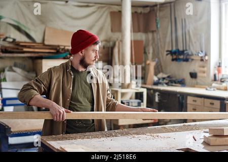 Jeune charpentier caucasien avec la barbe sur le visage portant la casquette tricotée rouge prenant la planche de bois pour couper et polir Banque D'Images