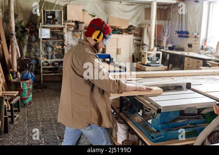 Menuisier professionnel portant des lunettes de sécurité et des écouteurs de protection coupant une planche en bois avec une scie à table Banque D'Images