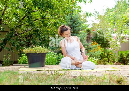 Une jeune femme s'assoit dans la position de lotus en aidant avec ses mains, pratique le yoga dans le jardin. Banque D'Images