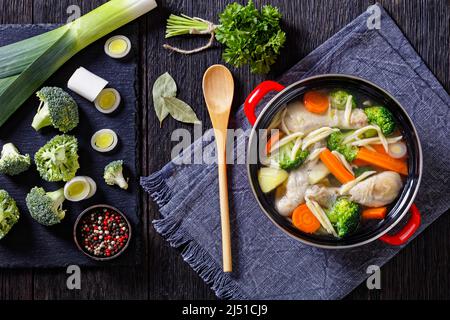 soupe de printemps au poulet avec brocoli, carottes, panais, poireaux et pâtes dans un pot rouge sur une table en bois sombre avec ingrédients sur un plateau en pierre noire, horizontal Banque D'Images