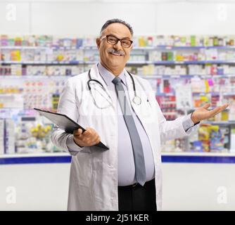 Pharmacien mûr souriant debout avec une planchette à pince dans une pharmacie et accueillant avec la main Banque D'Images