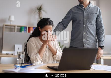 Une jeune femme agacée pleure près d'un ordinateur portable au bureau, une collègue l'aidant et la réconfortant Banque D'Images