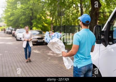 Jeune et professionnel liveur en uniforme bleu livre des fleurs à une femme cliente Banque D'Images