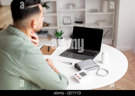 Jeune arabe dans la gorge de contrôles décontractés et regarde l'ordinateur portable avec écran vide dans le salon intérieur, vue sur l'épaule Banque D'Images