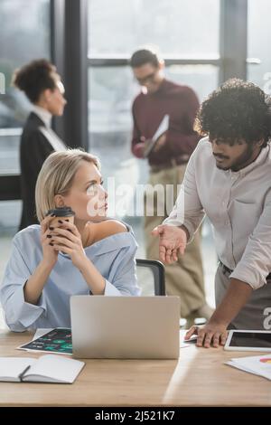 Homme d'affaires indien pointant vers un ordinateur portable près d'un collègue avec un café pour aller au bureau Banque D'Images