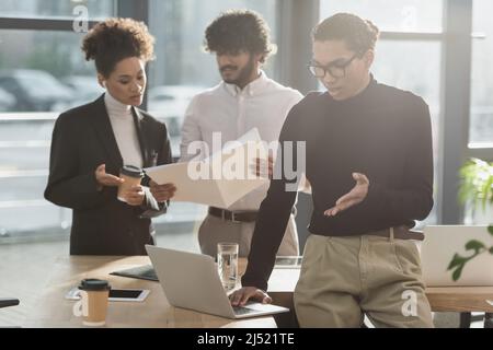 Un jeune homme d'affaires afro-américain ayant un chat vidéo sur ordinateur portable près de collègues interraciaux au bureau Banque D'Images