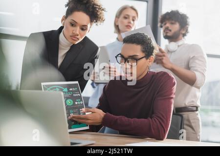 Un homme d'affaires afro-américain qui pointe sur le papier lors d'un appel vidéo sur un ordinateur portable, à proximité de collègues multiethniques flous au bureau Banque D'Images