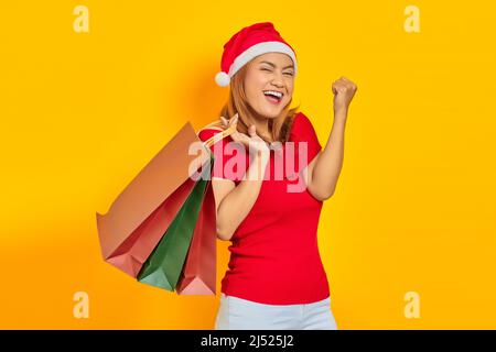 Jeune femme joyeuse en chapeau de Père Noël tenant des sacs de shopping et célébrant la satisfaction de shopping Banque D'Images