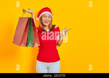 Jeune femme asiatique gaie dans le chapeau du Père Noël montrant des sacs d'achats et le doigt pointant vers l'espace de copie sur fond jaune Banque D'Images