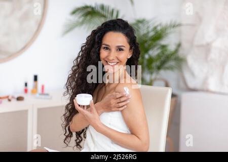 Jeune caucasienne souriante brune femme avec de longs cheveux bouclés dans une serviette applique de la crème sur l'épaule dans l'intérieur de la chambre Banque D'Images