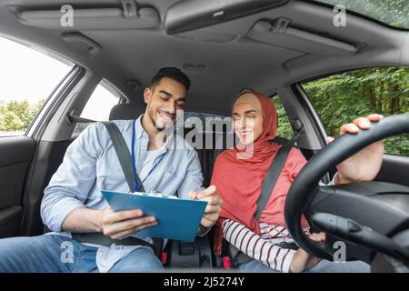 Un bon instructeur millennal de type musulman donne une excellente marque pour l'examen sur la route pour la femme dans le hijab au volant Banque D'Images