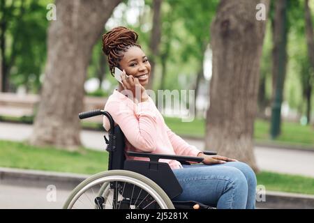 Joyeuse jeune Afro-américaine en fauteuil roulant parlant sur smartphone dans le parc urbain, espace libre Banque D'Images