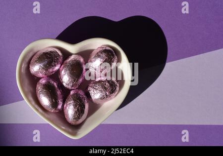 Œufs de Pâques au chocolat enveloppés d'une feuille dans un plat en forme de cœur isolé sur fond violet avec espace de copie Banque D'Images