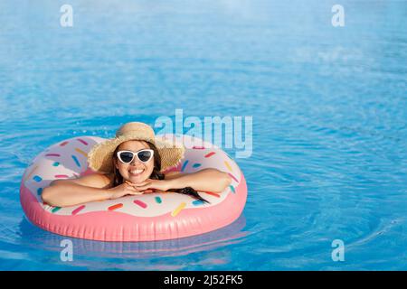 belle jeune femme en chapeau de paille et lunettes de soleil se détendant sur l'anneau gonflable dans la piscine Banque D'Images