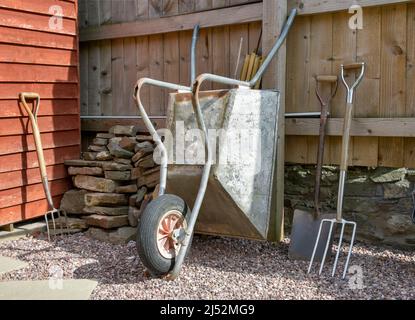 Coin jardin avec articles de jardinage dont brouette, grande bêche, grande fourche, pile de pierre, gants et bottes en caoutchouc contre la clôture en bois et le mur de pierre Banque D'Images