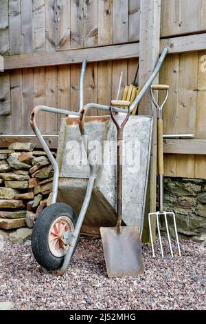 Articles de jardinage, y compris brouette, grande bêche, grande fourchette, gants et bottes en caoutchouc contre la clôture en bois et le mur en pierre Banque D'Images