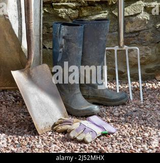 Articles de jardinage, y compris une grande pelle, une grande fourchette, des gants et des bottes en caoutchouc contre le mur de pierre Banque D'Images