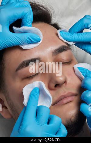 Un cosmétologue méconnaissable en gants médicaux qui retire les têtes noires du nez de l'homme d'âge moyen, à l'aide d'outils et d'un tampon en coton, vue latérale. Gros plan de la poignée Banque D'Images