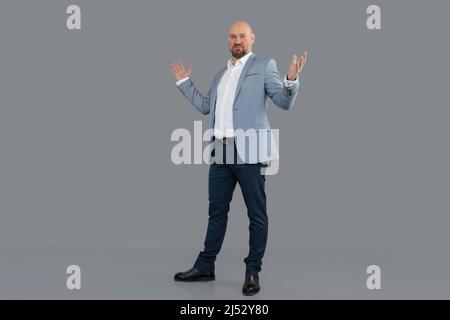 Portrait d'un beau chauve barbu d'âge moyen portant une veste bleue, un pantalon, une chemise blanche, debout, levant les mains. Banque D'Images