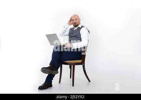 Portrait d'un homme d'âge moyen surpris avec gilet à carreaux gris, jeans bleus, chemise blanche, assis sur une chaise, tenant un ordinateur portable. Banque D'Images