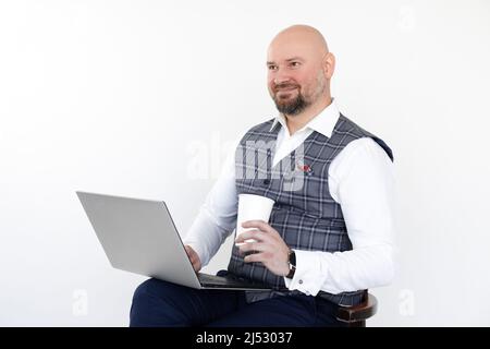 Portrait d'un homme heureux et attentionné avec gilet gris, Jean bleu, chemise blanche, ordinateur portable à porter assis et tasse de café à emporter. Banque D'Images