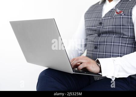 Photo courte d'homme d'affaires en gilet gris, jeans bleus, chemise blanche, assis sur une chaise, tenant un ordinateur portable, travail, saisie. Banque D'Images