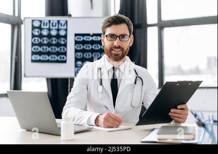 Fier médecin de médecine générale caucasienne avec des lunettes, thérapeute, en uniforme médical, assis sur le lieu de travail dans l'hôpital, utilise un ordinateur portable pour le travail, regarde la caméra, sourit. Médecine et concept de santé Banque D'Images