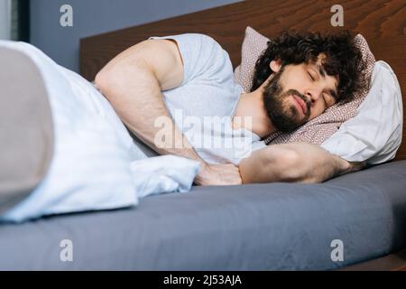 Vue rapprochée en angle bas du jeune homme barbu dormant paisiblement allongé sur le côté dans un grand lit confortable sous une couverture blanche Banque D'Images