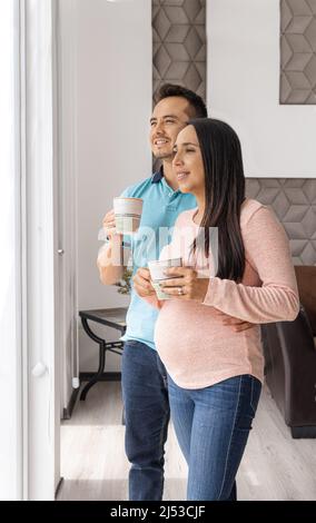 Photo verticale d'un homme souriant et de son partenaire enceinte qui a pris un verre dans leur appartement en regardant par la fenêtre Banque D'Images
